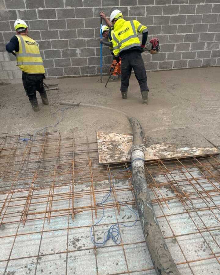 Construction workers in safety gear working on a concrete floor with a wall in the background.