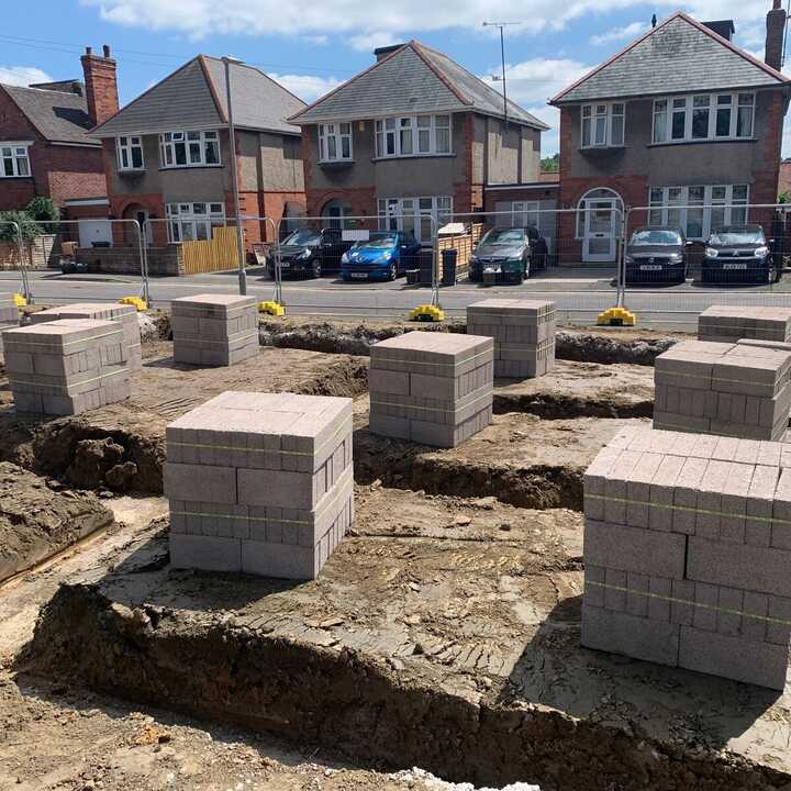 Construction site with stacked bricks on uneven ground and residential houses in the background.