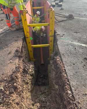 Construction workers near an open trench with a ladder and equipment visible.