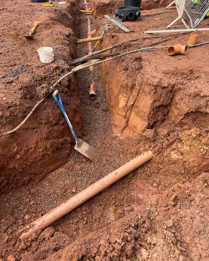 Excavation site showing a trench, tools, and exposed pipes in red earth.