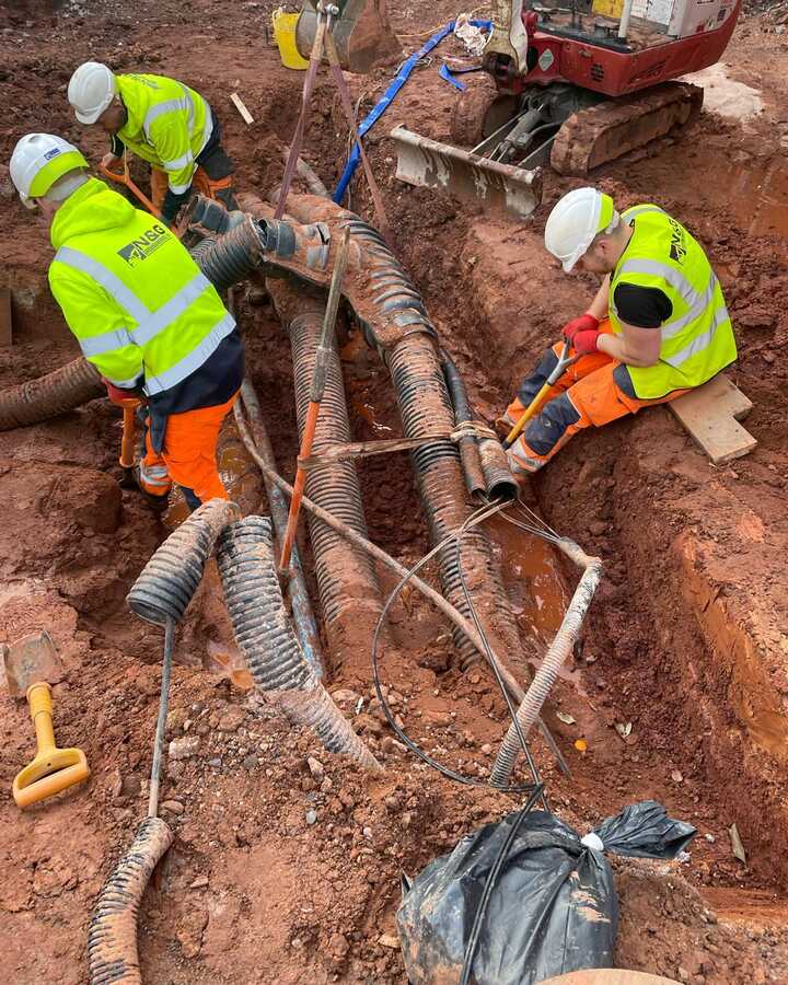 Workers in safety vests digging near exposed pipes in a trench.