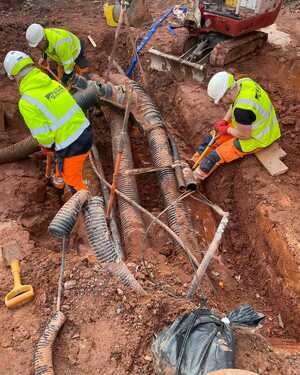 Workers in safety vests digging near exposed pipes in a trench.