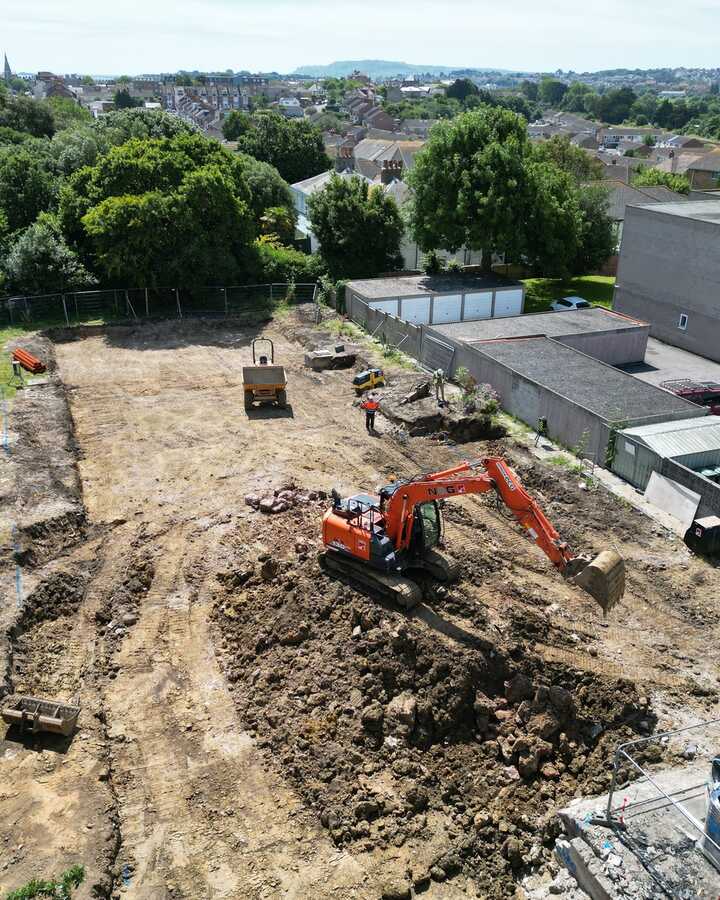 Construction site with an excavator and cleared land surrounded by buildings and trees.