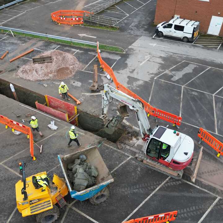 Construction site with workers, excavator, and trucks near road barriers and parked vehicles.