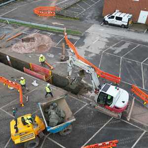 Construction site with workers, excavator, and trucks near road barriers and parked vehicles.