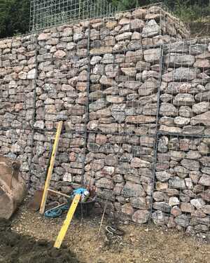 Gabion stone wall construction with tools and materials nearby on a sloped area.