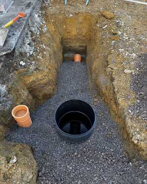 Excavated trench with gravel, black drainage barrel, and orange pipe.