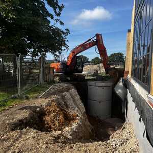 Excavator digging near a building, with a pile of dirt and a barrel visible.